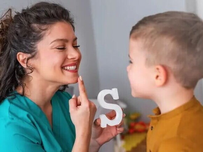 Uma mulher sorrindo, apontando para a própria boca e segurando um objeto em forma de letra “S”, enquanto uma criança observa e tenta imitar a ação da mulher. Uma mulher sorrindo, apontando para a própria boca e segurando um objeto em forma de letra “S”, enquanto uma criança observa e tenta imitar a ação da mulher.