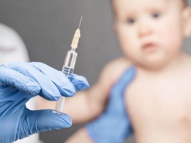 Mão com luva cirúrgica segurando seringa com a vacina da coqueluche, enquanto bebê aguarda a aplicação