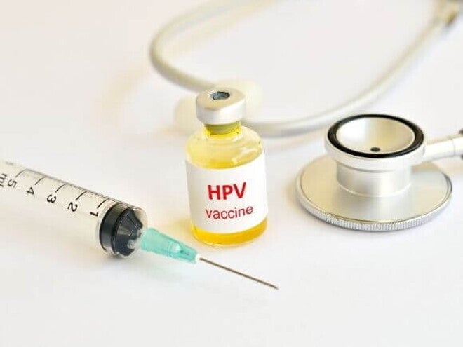 Vista superior de um frasco de vacina rotulado "HPV vaccine" com líquido amarelado, colocado entre uma seringa hipodérmica e um estetoscópio médico, todos dispostos sobre uma superfície branca.