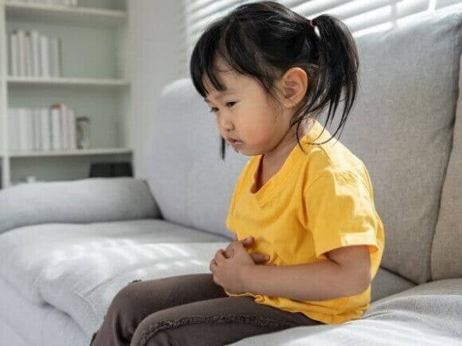 Uma criança sentada no sofá com a mão na barriga, expressando dor. A imagem representa o mal-estar causado por verme infantil.