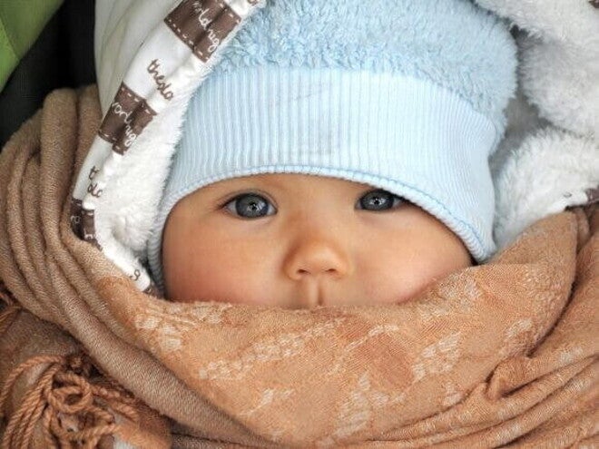 Um close-up de um bebê usando um chapéu azul e um cobertor marrom enrolado em volta do rosto, mostrando como agasalhar um bebê para o frio e como vestir um bebê de acordo com a temperatura.