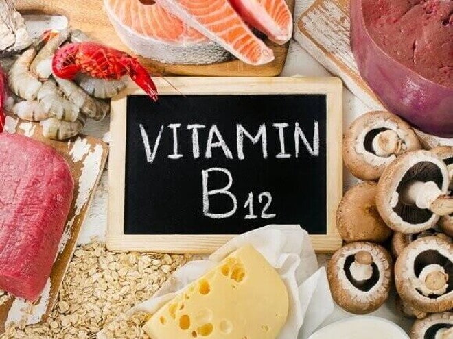 Uma composição de alimentos ricos em vitamina B12, com um pequeno quadro negro no centro com a inscrição "Vitamina B12" em giz. 