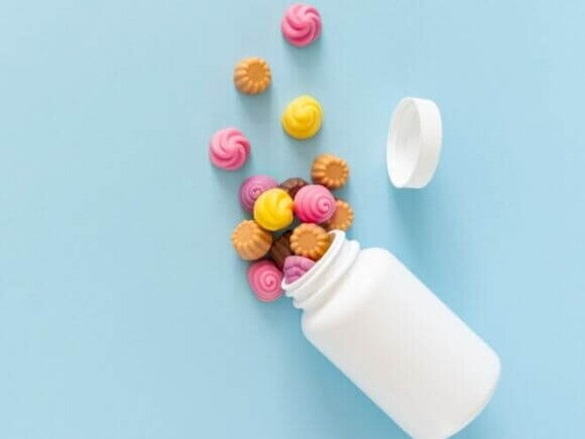 Vitaminas para criança Visão de cima de gomas de vitaminas coloridas, em formato de espiral ou flor, que caíram de um pote branco sem rótulo, sobre uma superfície azul clara.