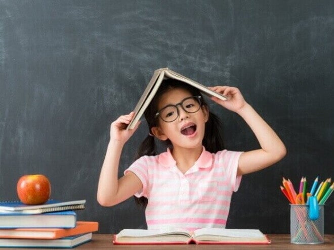 Uma menina com óculos, sentada à mesa, faz uma pose divertida com um livro aberto sobre a cabeça. Ela sorri com a boca aberta em frente a um quadro negro, com um monte de livros e lápis de cor ao seu lado. A imagem é lúdica e representa a criatividade e a diversão do aprendizado, remetendo ao clima de volta às aulas.