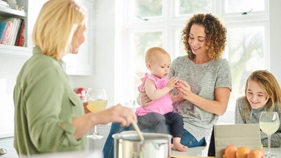 Uma mulher em um ambiente doméstico sorrindo e segurando um bebê no colo ao lado de uma menina, enquanto outra mulher está mexendo em uma panela. Uma mulher em um ambiente doméstico sorrindo e segurando um bebê no colo ao lado de uma menina, enquanto outra mulher está mexendo em uma panela.