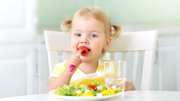Criança sorrindo, sentada à mesa e comendo uma salada saudável, com um copo de água ao lado. A imagem sugere que uma boa alimentação e hidratação podem ajudar a aliviar dor de cabeça em criança, sendo uma das medidas preventivas.