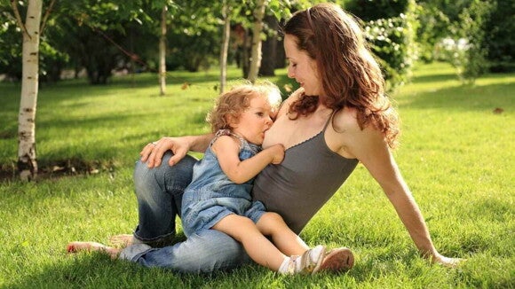 Mãe sentada no gramado ao ar livre amamentando sua filha pequena (cerca de 2 anos), que está sentada em seu colo.