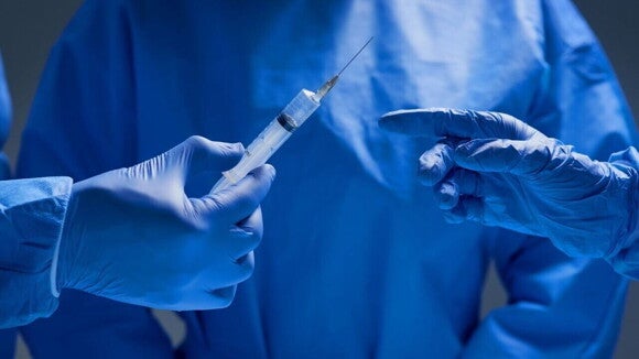 Close-up de duas mãos enluvadas, uma segurando uma seringa com agulha e a outra apontando.