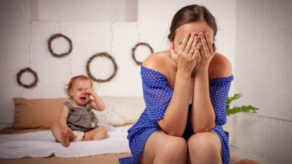 Uma mulher sentada na cama com as mãos cobrindo o rosto, enquanto um bebê está sentado na cama atrás dela, chorando. Uma mulher sentada na cama com as mãos cobrindo o rosto, enquanto um bebê está sentado na cama atrás dela, chorando.