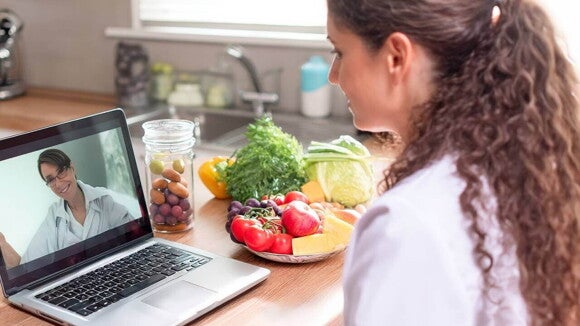 Mulher faz uma consulta online com um profissional de saúde sobre dietas de exclusão para APLV, com diversas frutas e vegetais sobre a mesa, representando a alimentação durante a amamentação.