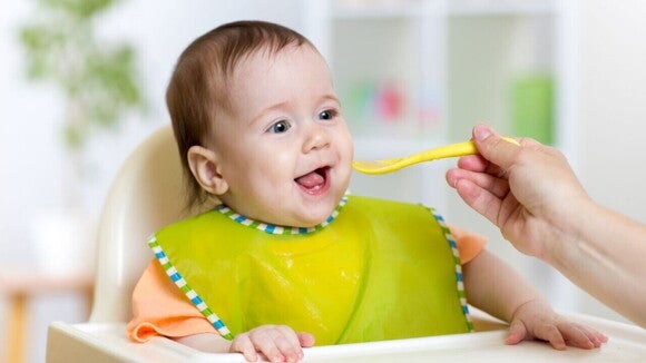 Um bebê sorrindo enquanto é alimentado com uma colher. A imagem pode ser usada para ilustrar a introdução alimentar e a importância de identificar alergias como a APLV em bebê.