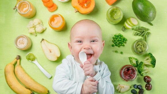 Um bebê sorridente, deitado em um fundo verde, segurando uma colher. Ele está cercado por várias frutas e vegetais e potes de papinhas de cores diversas, simbolizando a alimentação complementar e a variedade de alimentos livres de leite.
