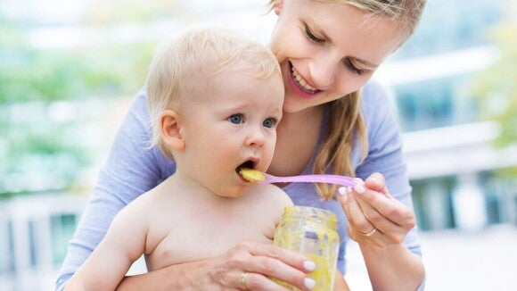 Uma mãe sorridente alimenta um bebê, com uma colher de papinha, destacando o cuidado com a dieta e a alimentação infantil.