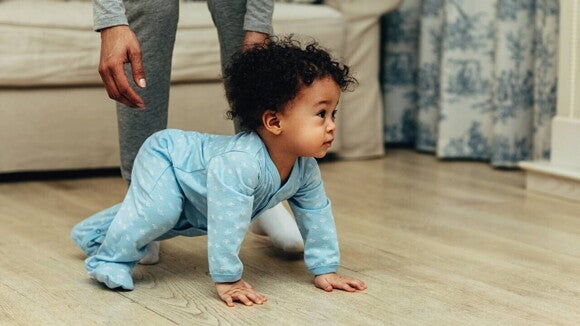 Um bebê engatinhando em um chão de madeira clara. Ele veste um macacão azul claro com estampas brancas e parece estar olhando para a direita. Ao fundo, as pernas de um adulto em calça cinza estão visíveis, sugerindo que o adulto está por perto. Um bebê engatinhando em um chão de madeira clara. Ele veste um macacão azul claro com estampas brancas e parece estar olhando para a direita. Ao fundo, as pernas de um adulto em calça cinza estão visíveis, sugerindo que o adulto está por perto.