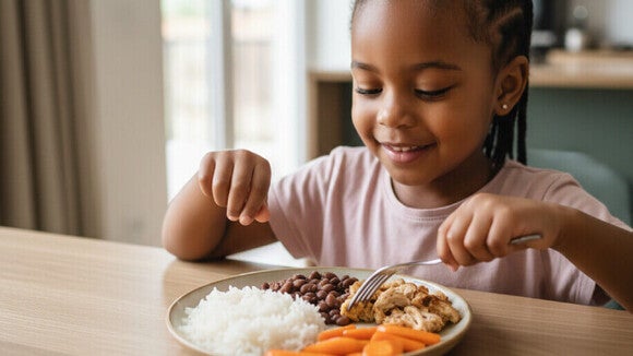Criança feliz comendo prato colorido de arroz, feijão, frango e cenoura em ambiente acolhedor.