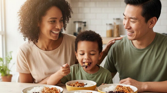 Família diversa almoçando arroz, feijão, frango desfiado e cenoura cozida em cozinha iluminada.