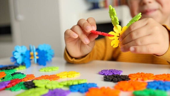 Um close-up mostra as mãos de uma criança montando um brinquedo de encaixe colorido e com formato de flor. Diversas outras peças do brinquedo estão espalhadas pela mesa branca, destacando a interação e o foco da criança na atividade de construção, ilustrando o conceito de brincadeiras sensoriais. Um close-up mostra as mãos de uma criança montando um brinquedo de encaixe colorido e com formato de flor. Diversas outras peças do brinquedo estão espalhadas pela mesa branca, destacando a interação e o foco da criança na atividade de construção, ilustrando o conceito de brincadeiras sensoriais.