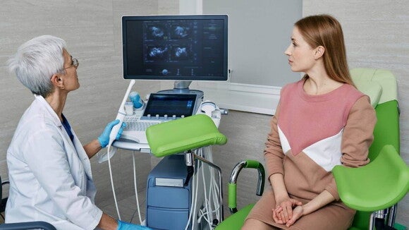 Mulher em uma cadeira de exame ginecológico ou de ultrassom conversando com uma médica, com uma tela de ultrassom visível ao fundo.