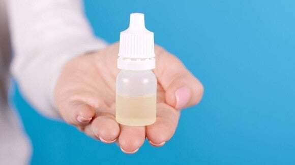 Uma mão estende-se para a câmera, segurando um pequeno frasco branco e translúcido de colírio contra um fundo azul sólido.