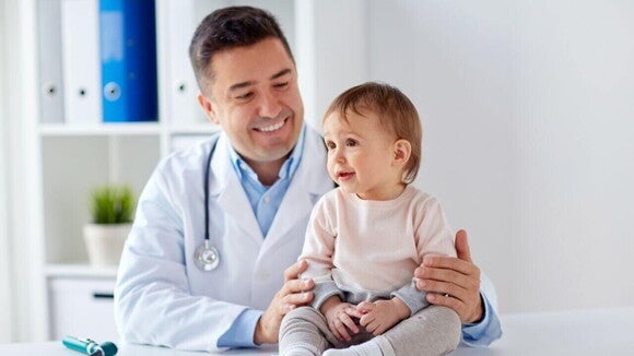 Com quantos meses o bebê pode tomar chá Um pediatra sorridente em um consultório, vestindo um jaleco branco e estetoscópio, segura um bebê que está sentado na mesa de exame.