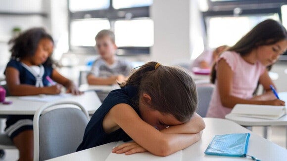Uma menina sentada em uma sala de aula com a cabeça apoiada nos braços sobre a mesa, demonstrando desânimo ou dificuldade de concentração. Ao fundo, outros alunos estudam.
