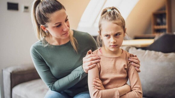Uma mãe está sentada em um sofá, abraçando uma menina com uma expressão emburrada e braços cruzados. A mãe olha para a filha com preocupação e ternura, enquanto tenta confortá-la. A cena ilustra uma interação entre mãe e filha durante um momento de frustração da criança, representando uma abordagem de como trabalhar essa emoção. Uma mãe está sentada em um sofá, abraçando uma menina com uma expressão emburrada e braços cruzados. A mãe olha para a filha com preocupação e ternura, enquanto tenta confortá-la. A cena ilustra uma interação entre mãe e filha durante um momento de frustração da criança, representando uma abordagem de como trabalhar essa emoção.