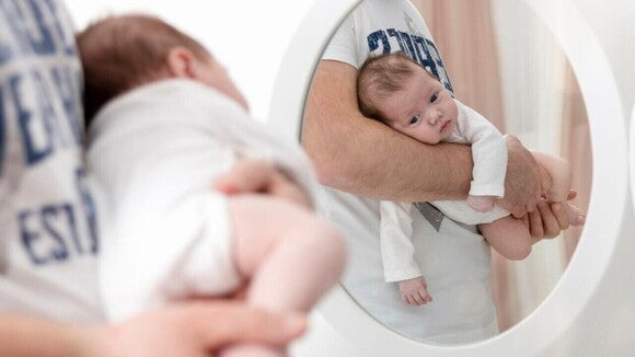 Um pai segura um bebê recém-nascido de bruços e o posiciona na frente de um espelho redondo.
