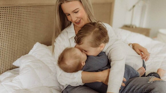 Uma mãe está sentada em uma cama, segurando seus dois filhos. O filho mais velho abraça o irmão mais novo com carinho, enquanto a mãe observa a interação com um sorriso. A cena de afeto familiar destaca a adaptação e o vínculo construído entre os irmãos, referindo-se à palavra-chave mudança de comportamento com a chegada do irmão. Uma mãe está sentada em uma cama, segurando seus dois filhos. O filho mais velho abraça o irmão mais novo com carinho, enquanto a mãe observa a interação com um sorriso. A cena de afeto familiar destaca a adaptação e o vínculo construído entre os irmãos, referindo-se à palavra-chave mudança de comportamento com a chegada do irmão.