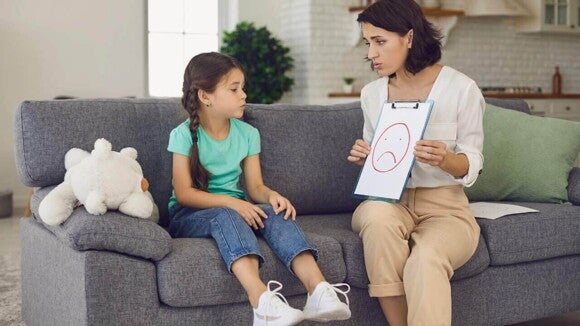 Mulher adulta sentada em um sofá conversando seriamente com uma menina pré-adolescente, segurando uma prancheta com um rosto triste desenhado.