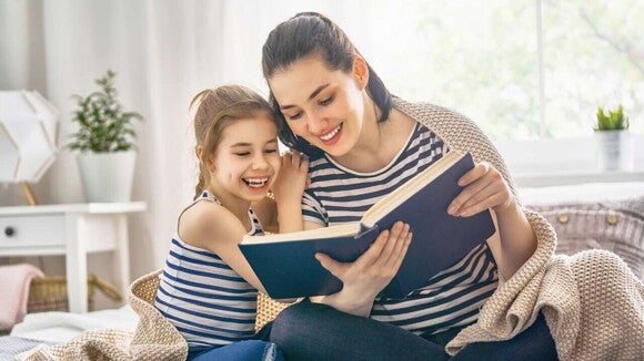 Uma mãe e sua filha pequena estão sentadas em um sofá ou cama. A mãe segura um livro aberto e sorri enquanto a filha, ao seu lado, ri alegremente, olhando para o livro.
