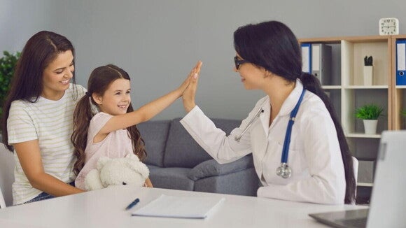 Uma criança sentada no colo de uma mulher, fazendo um toque de mão com uma médica sentada à sua frente Uma criança sentada no colo de uma mulher, fazendo um toque de mão com uma médica sentada à sua frente