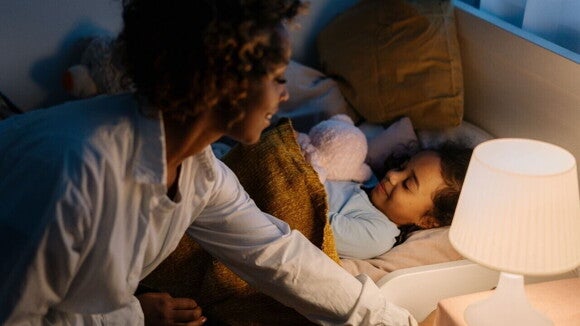 Mãe se abaixando para apagar um abajur aceso, com a filha pequena na cama ao lado, ajudando a acalmar a criança antes de dormir.