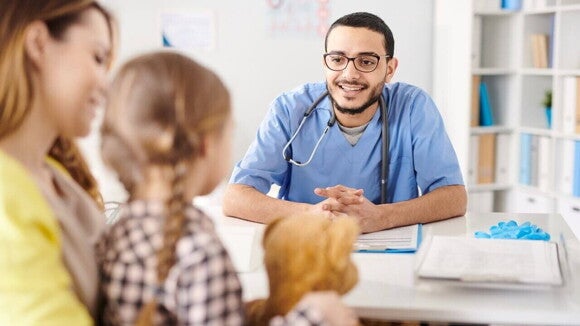 Vista das costas de uma criança acompanhada pela mãe durante uma consulta médica. À frente delas, um pediatra sorridente de óculos e uniforme azul tenta transmitir confiança. A menina segura um urso de pelúcia.