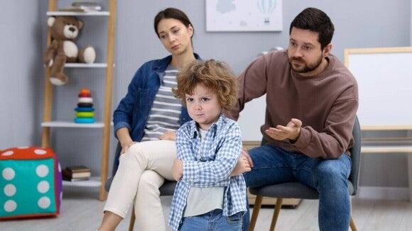 Um menino com os braços cruzados e expressão de frustração está à frente de seus pais, que parecem preocupados e tentando lidar com o comportamento, ilustrando uma crise de raiva ou birra.