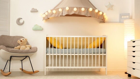 Um quarto de bebê moderno e aconchegante com paredes brancas e iluminação suave. Um berço branco de madeira está no centro, com travesseiros amarelos e um lençol cinza.
