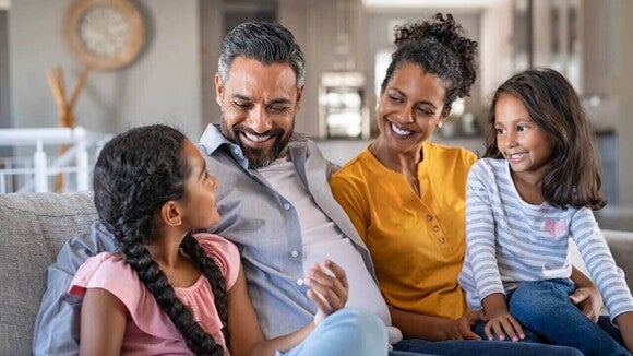 Uma família de quatro pessoas está sentada em um sofá na sala de estar. A mãe e o pai estão sorrindo e olhando para suas duas filhas. Uma família de quatro pessoas está sentada em um sofá na sala de estar. A mãe e o pai estão sorrindo e olhando para suas duas filhas.