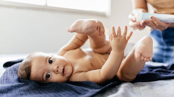 Um bebê deitado segurando o pé, enquanto uma pessoa manipula um frasco de creme. Um bebê deitado segurando o pé, enquanto uma pessoa manipula um frasco de creme.