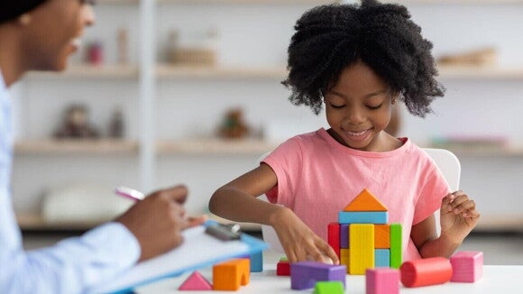 Terapeuta de desenvolvimento infantil observa uma criança sorridente, montando uma torre com blocos coloridos, em um ambiente de consultório. A cena ilustra atividades que estimulam o desenvolvimento cerebral infantil através do brincar e da interação.