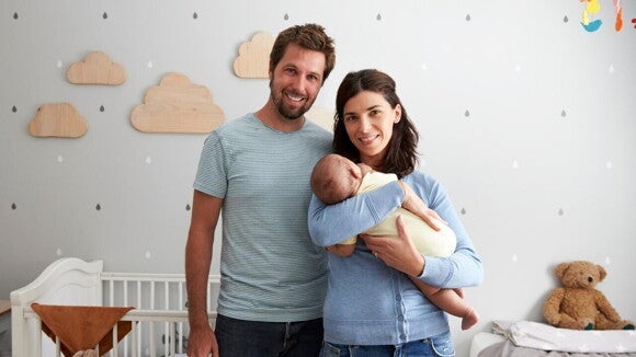 Um homem e uma mulher em um quarto infantil, sorrindo para a câmera, com a mulher segurando um bebê nos braços. Um homem e uma mulher em um quarto infantil, sorrindo para a câmera, com a mulher segurando um bebê nos braços.