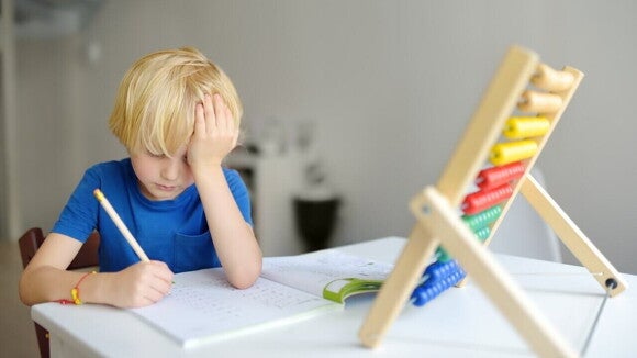 Criança loira expressando frustração e dificuldade de aprendizagem enquanto tenta resolver exercícios de matemática com o auxílio de um ábaco sobre a mesa.