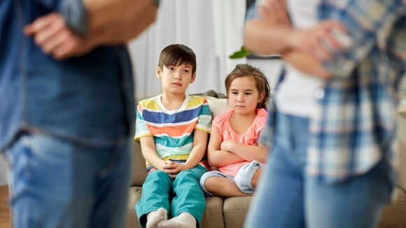 Duas crianças sentadas em um sofá com expressões tristes, enquanto seus pais discutem em primeiro plano, representando o impacto emocional do divórcio nos filhos. Duas crianças sentadas em um sofá com expressões tristes, enquanto seus pais discutem em primeiro plano, representando o impacto emocional do divórcio nos filhos.