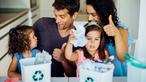 Pais e suas duas filhas participam de uma atividade de reciclagem em casa, com as crianças colocando papel amassado em lixeiras com o símbolo de reciclagem, enquanto os pais incentivam. Pais e suas duas filhas participam de uma atividade de reciclagem em casa, com as crianças colocando papel amassado em lixeiras com o símbolo de reciclagem, enquanto os pais incentivam.