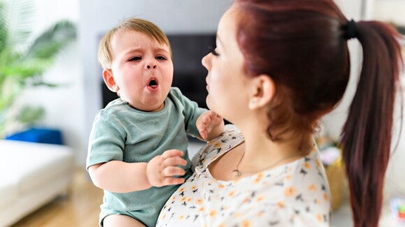 Uma mãe segura um bebê chorando, com a boca aberta e as mãos levantadas, sugerindo possível desconforto ou um engasgo. Uma mãe segura um bebê chorando, com a boca aberta e as mãos levantadas, sugerindo possível desconforto ou um engasgo.