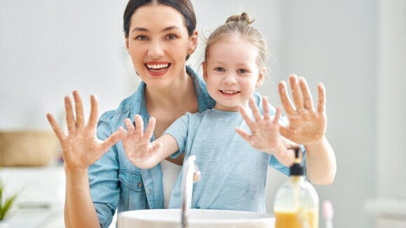 Uma mulher e uma menina sorrindo, mostrando as mãos ensaboadas, em frente a uma pia. Uma mulher e uma menina sorrindo, mostrando as mãos ensaboadas, em frente a uma pia.