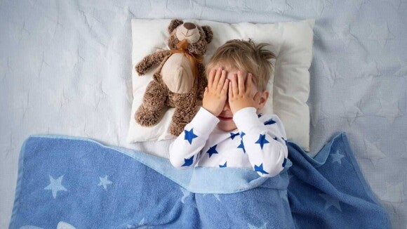 Criança deitada na cama ao lado de um urso de pelúcia, cobrindo o rosto com as mãos, usando pijama com estrelas azuis e cobertor azul com estrelas. Criança deitada na cama ao lado de um urso de pelúcia, cobrindo o rosto com as mãos, usando pijama com estrelas azuis e cobertor azul com estrelas.