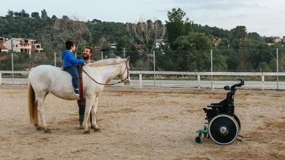 Criança montada em um cavalo ao lado de um homem, com uma cadeira de rodas à frent Criança montada em um cavalo ao lado de um homem, com uma cadeira de rodas à frent