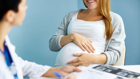 Uma mulher grávida sorri durante uma consulta médica, segurando a barriga, com imagens de ultrassom sobre a mesa. A imagem ilustra o acompanhamento pré-natal e o diagnóstico de condições como a espinha bífida.