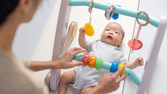 Um bebê deitado de costas em um tapete de atividades, enquanto uma pessoa manipula um brinquedo colorido pendurado em uma estrutura acima do bebê. Um bebê deitado de costas em um tapete de atividades, enquanto uma pessoa manipula um brinquedo colorido pendurado em uma estrutura acima do bebê.
