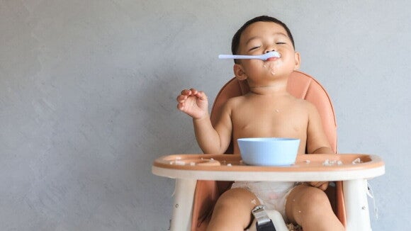 Uma criança sentada em uma cadeira de alimentação, suja de comida, com uma colher na boca, parecendo feliz após uma refeição.