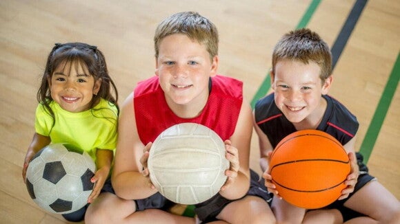 Crianças sentadas e sorrindo, segurando bolas de modalidades esportivas diferentes.